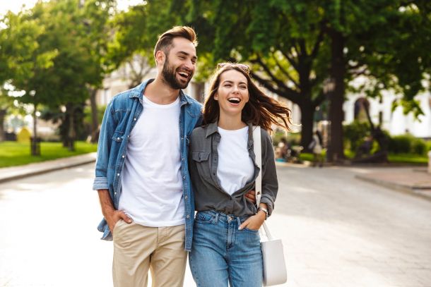 A smiling couple walks outdoors on a sunny day, dressed casually in jeans and light shirts, with trees and greenery in the background.
